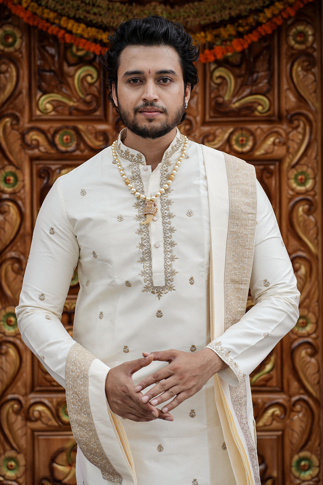 Man in traditional white sherwani with gold embroidery against a decorative wooden background
