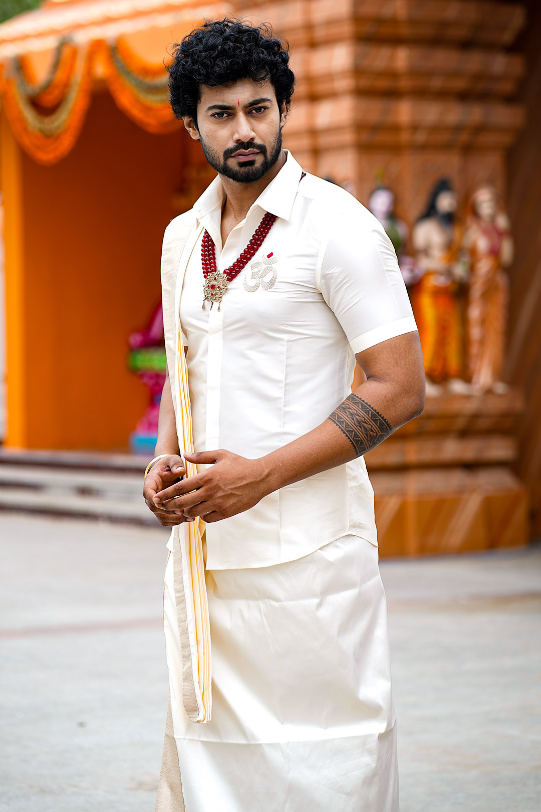 Man in traditional white outfit holding a staff in front of an orange backdrop with statues.