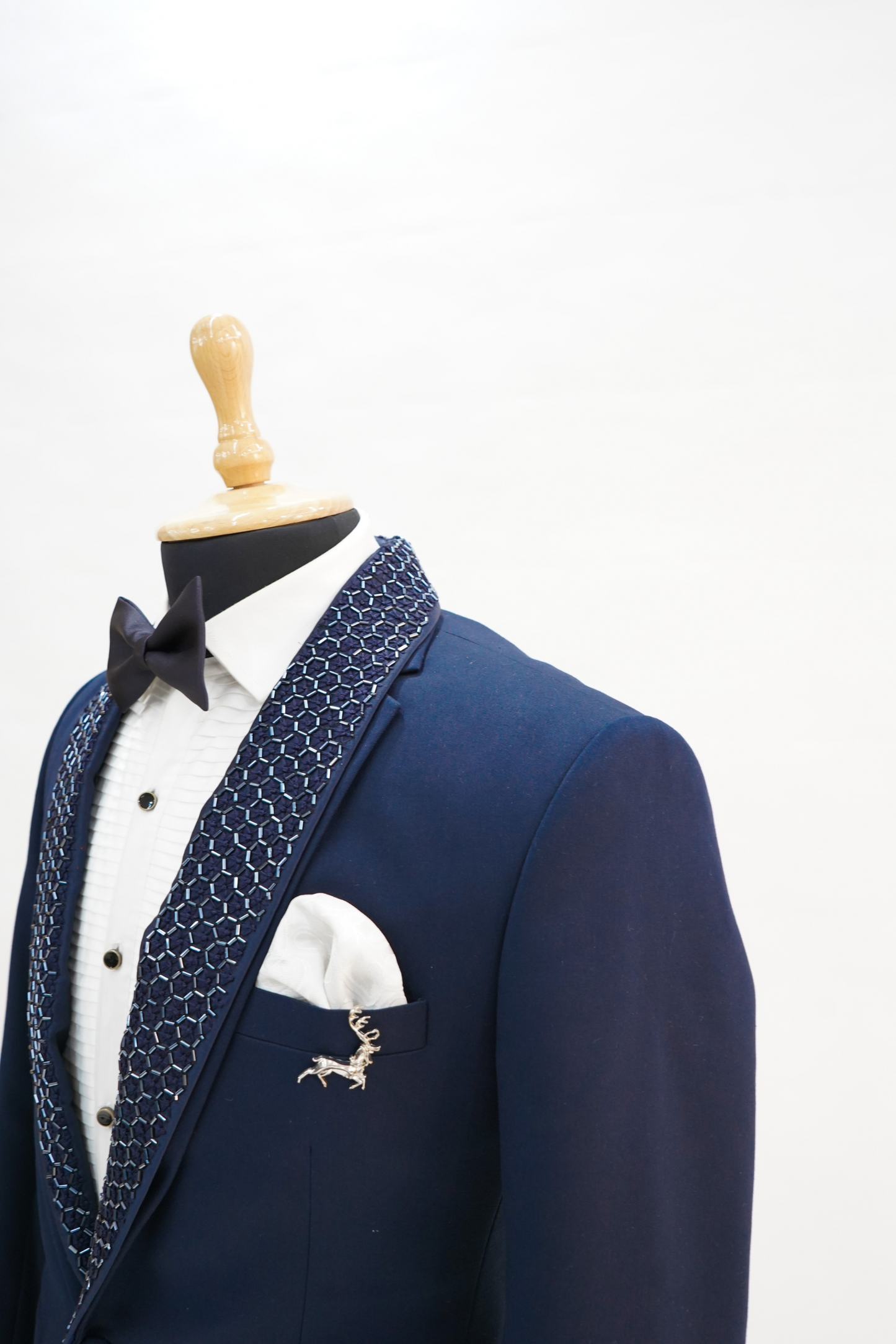 A man sitting on a yellow chair wearing a navy blue wool crepe 2-piece tuxedo with a detachable lapel, consisting of a jacket and trousers, accessorized with a bow tie and a handkerchief.