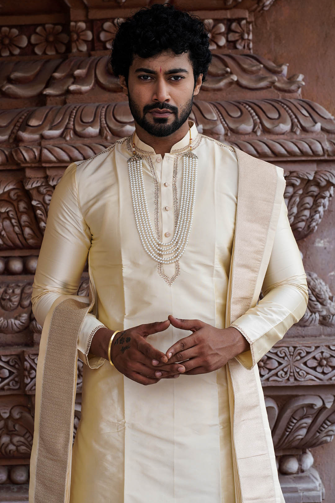 Man wearing a traditional cream sherwani with intricate patterns in the background