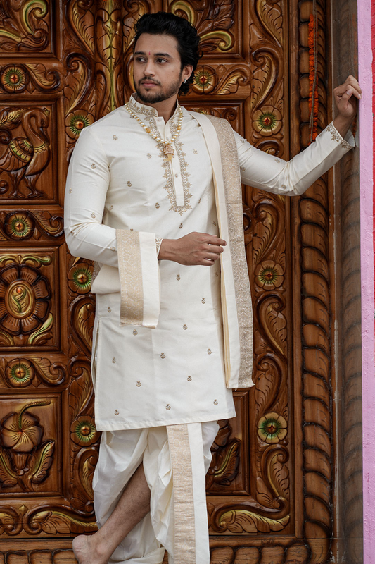 Man in traditional white sherwani with gold embroidery against a decorative wooden background