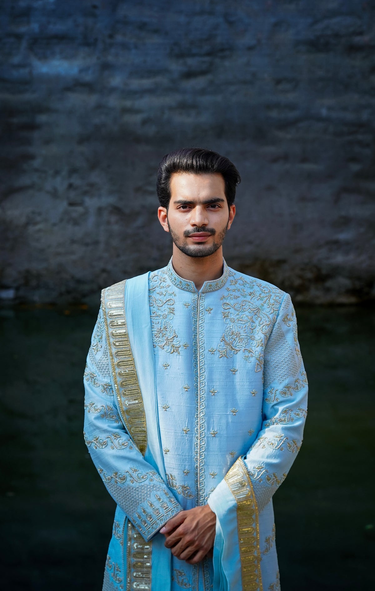 A man standing by a water body, wearing a traditional Sherwani with blue and gold colors, featuring a dupion silk dhoti and khandwa.