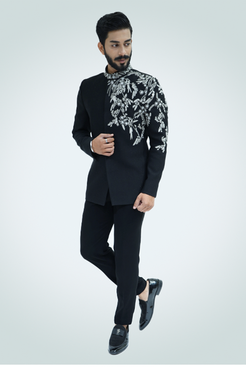 A man wearing a black suit with silver embroidery standing against a light background.