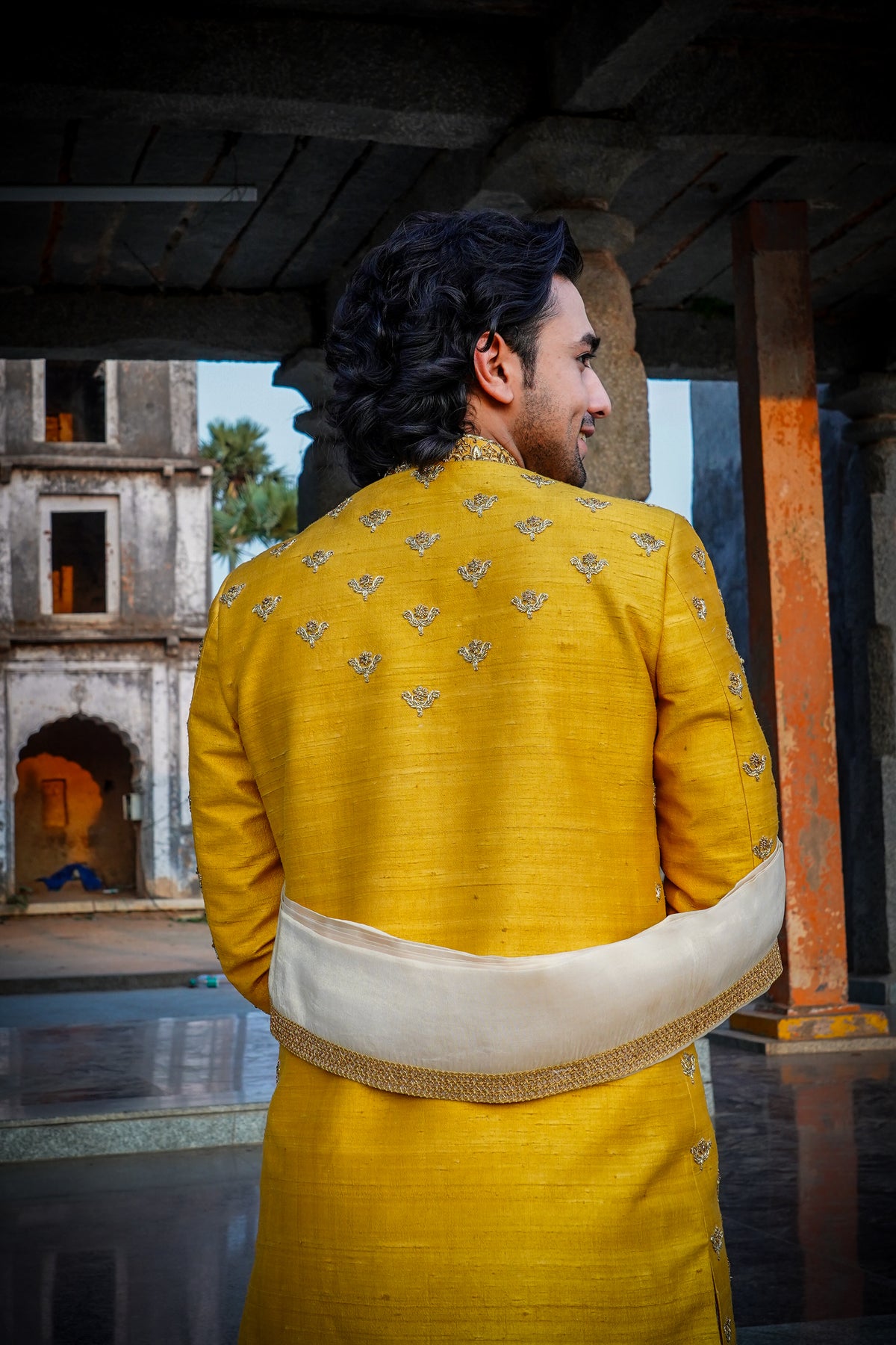 A man wearing a yellow sherwani with gold embroidery and a turban, standing in a traditional setting with a textured background.