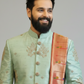 Man wearing a light green embroidered sherwani with a pink and gold sash on a gray background