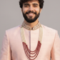 Man wearing a light pink traditional outfit with a necklace against a gray background