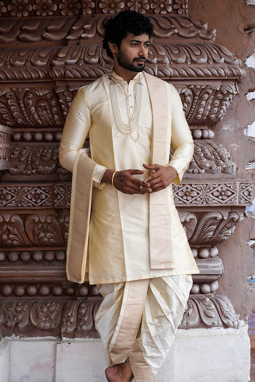Man wearing a cream traditional outfit standing against an ornate stone wall.