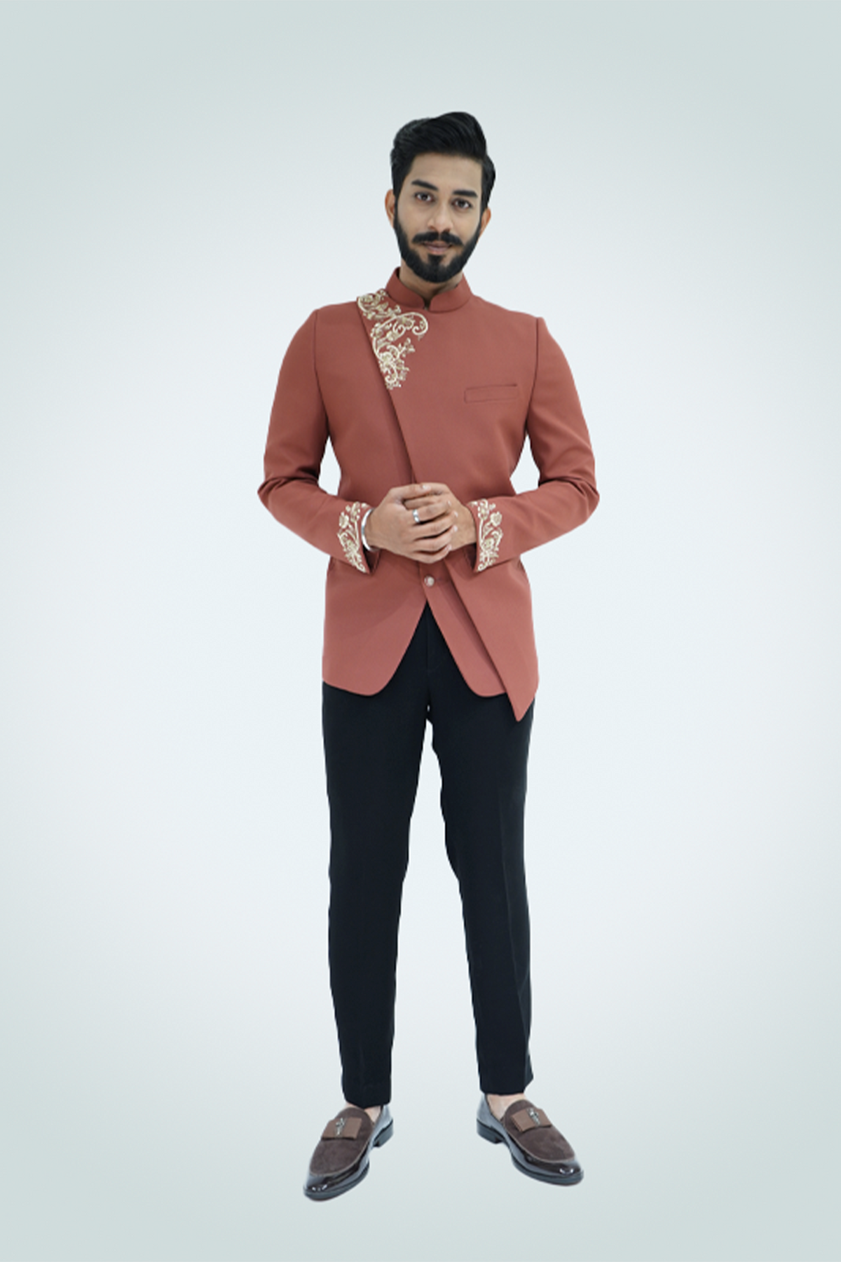 A man wearing a rust-colored Jodhpuri suit with gold embroidery, featuring a mandarin collar and cross cut design.