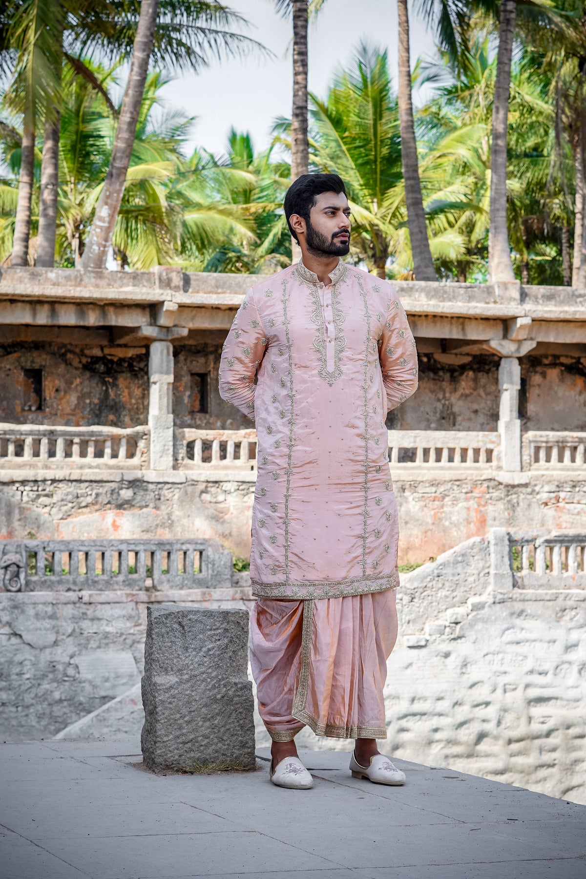 A man standing by a water body, wearing a traditional Sherwani with blue and gold colors, featuring a dupion silk dhoti and khandwa.