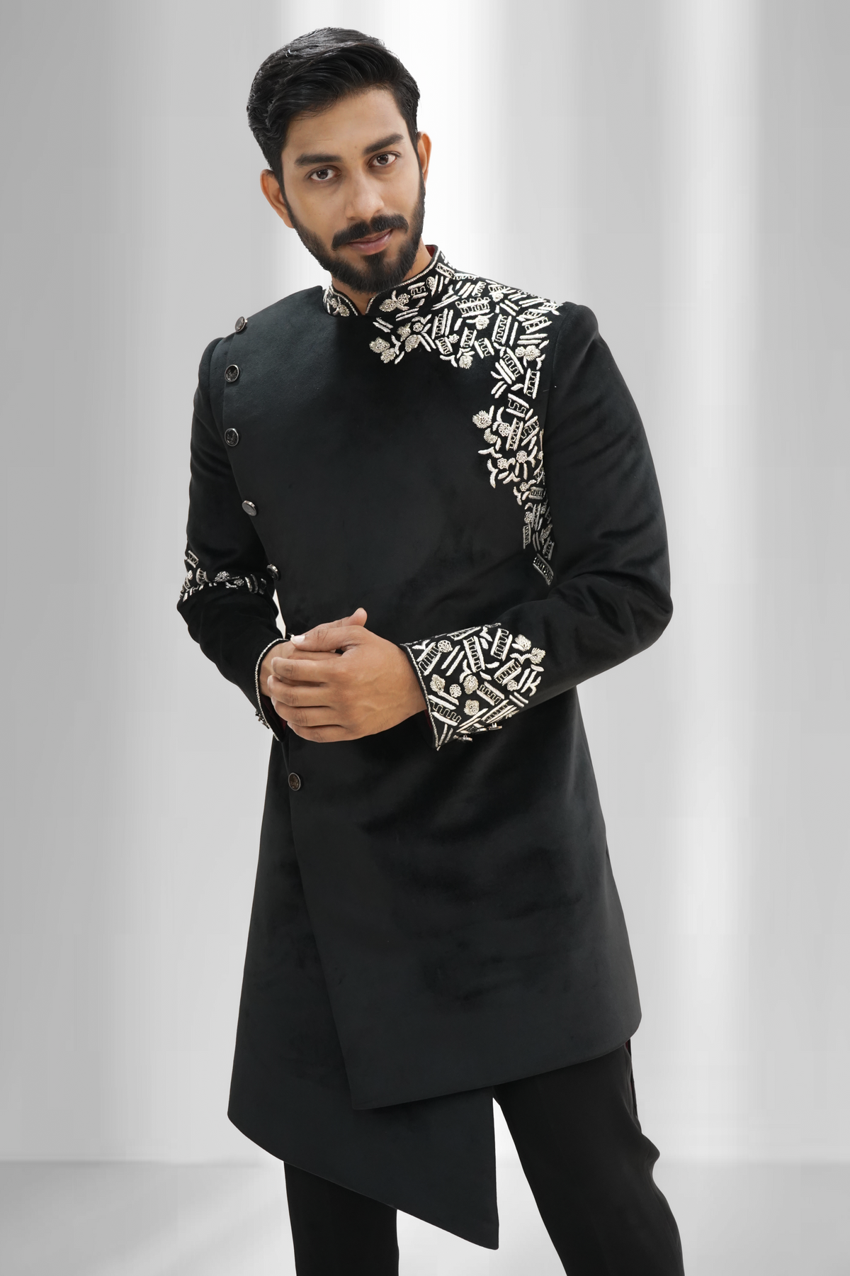 A man wearing a black asymmetrical Indo-Western sherwani with silver zardozi work on the sleeves and chest.