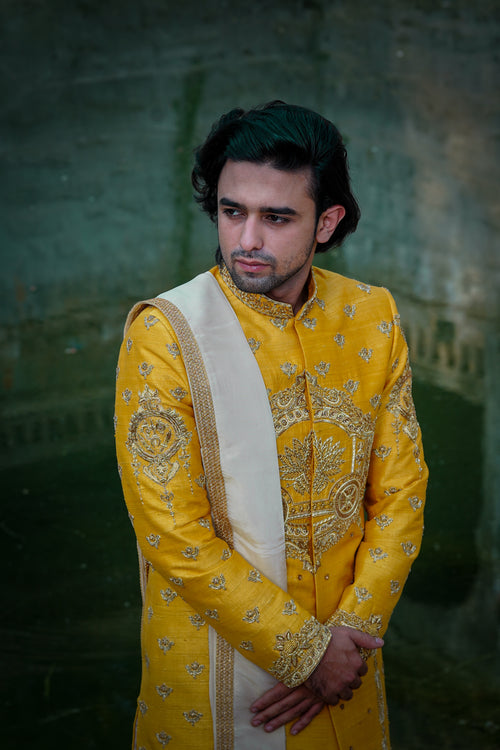 A man wearing a yellow sherwani with gold embroidery and a turban, standing in a traditional setting with a textured background.