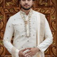 Man in traditional white sherwani with gold embroidery against a decorative wooden background