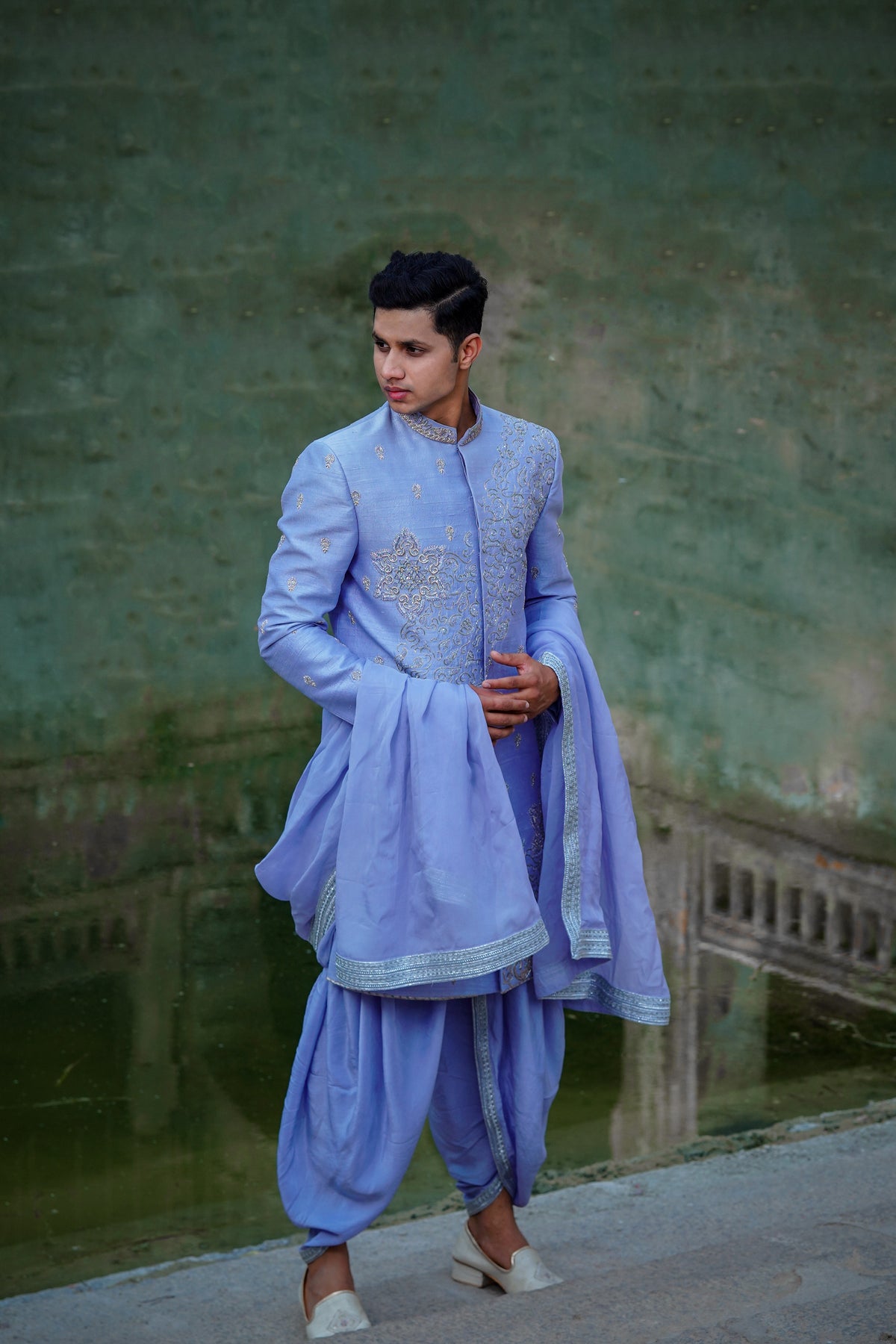 A man wearing a blue Dupam Sherwani with intricate embellishments and opulent embroidery, standing against an outdoor backdrop.