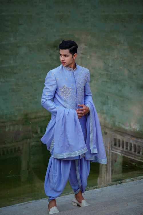 A man wearing a blue Dupam Sherwani with intricate embellishments and opulent embroidery, standing against an outdoor backdrop.