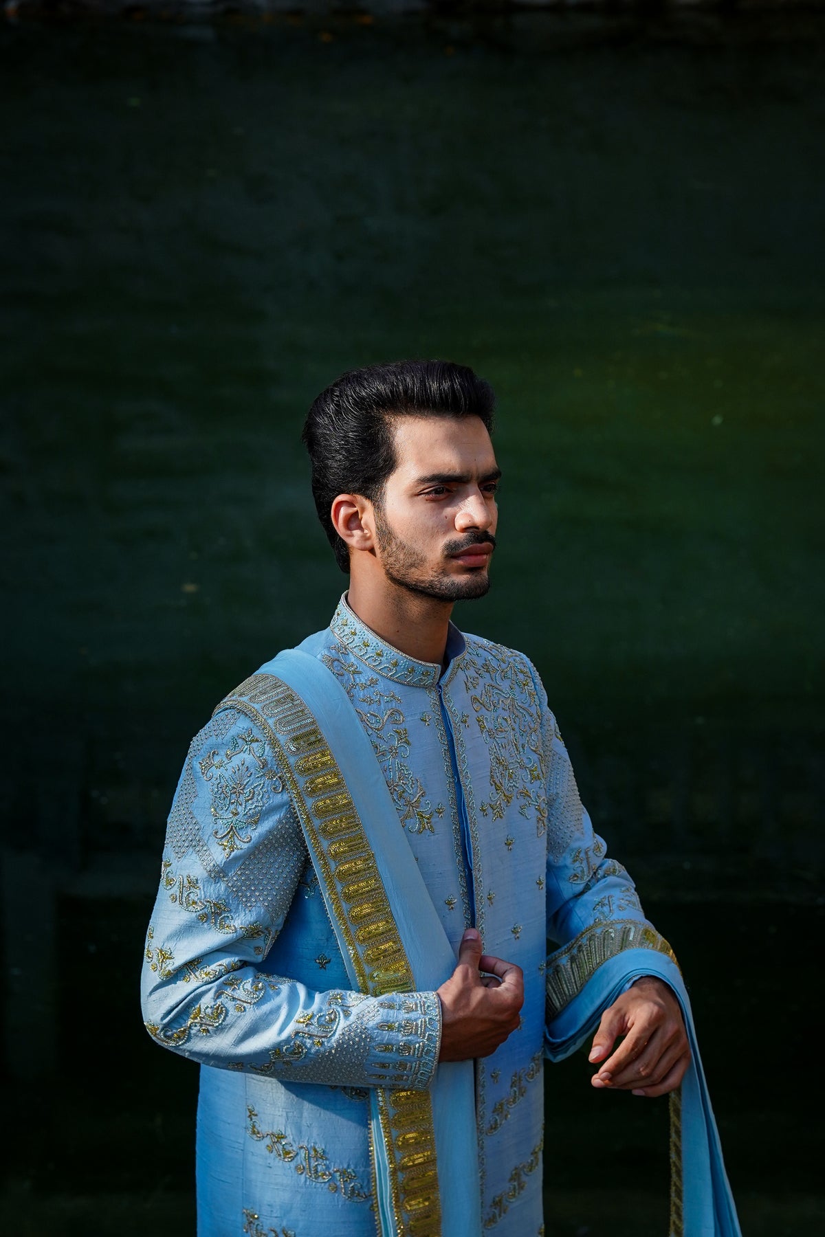 A man standing by a water body, wearing a traditional Sherwani with blue and gold colors, featuring a dupion silk dhoti and khandwa.