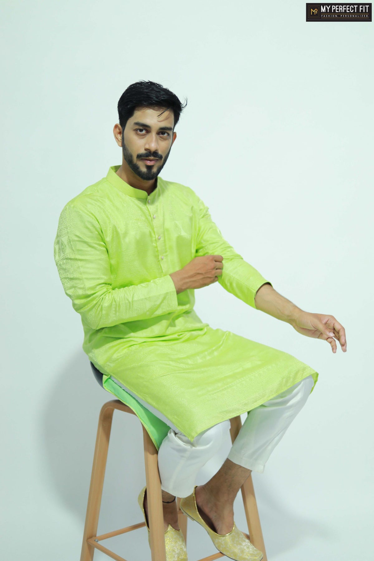 A person seating in a chair  a white background, wearing a green jacquard kurta with full-length sleeves, paired with white trousers.