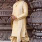 Man wearing a traditional cream-colored sherwani with a gold border against an ornate architectural background.