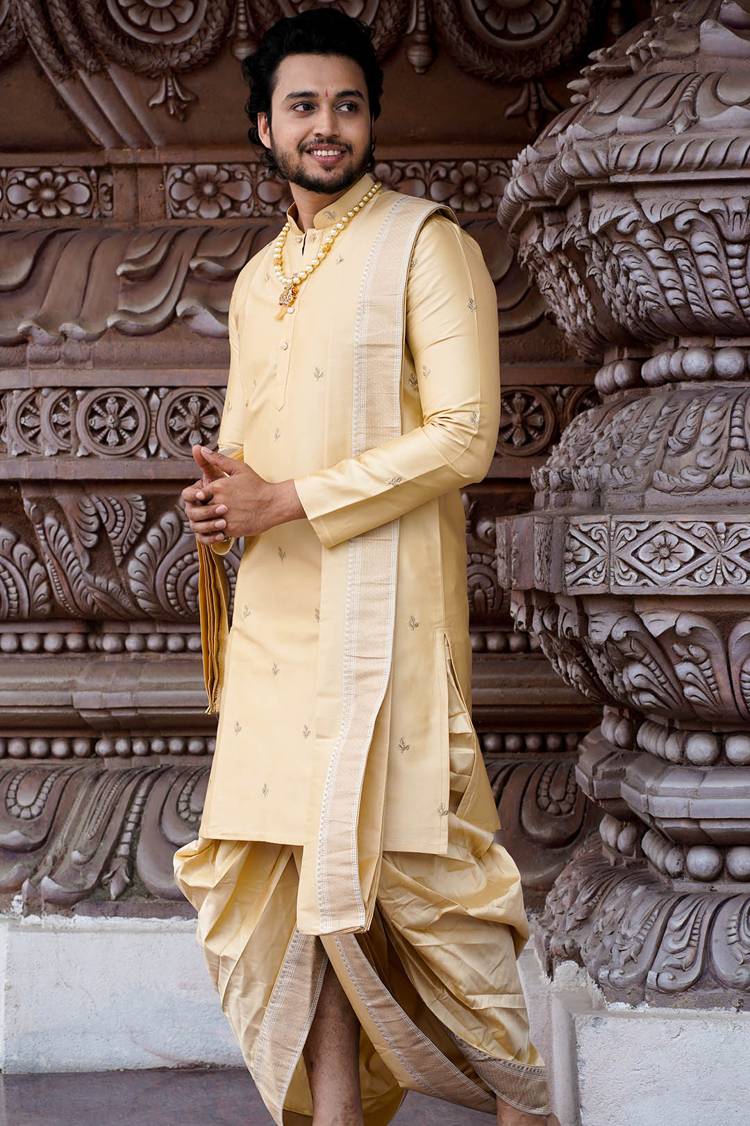 Man wearing a traditional cream-colored sherwani with a gold border against an ornate architectural background.