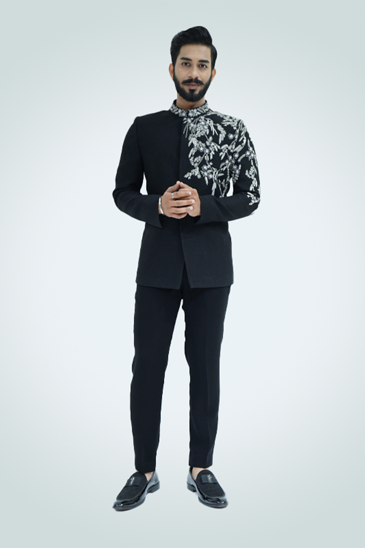 A man wearing a black suit with silver embroidery standing against a light background.