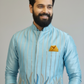 Man wearing a light blue traditional outfit with a yellow pocket square against a gray background