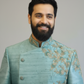 Man wearing a light blue embroidered traditional outfit against a plain background