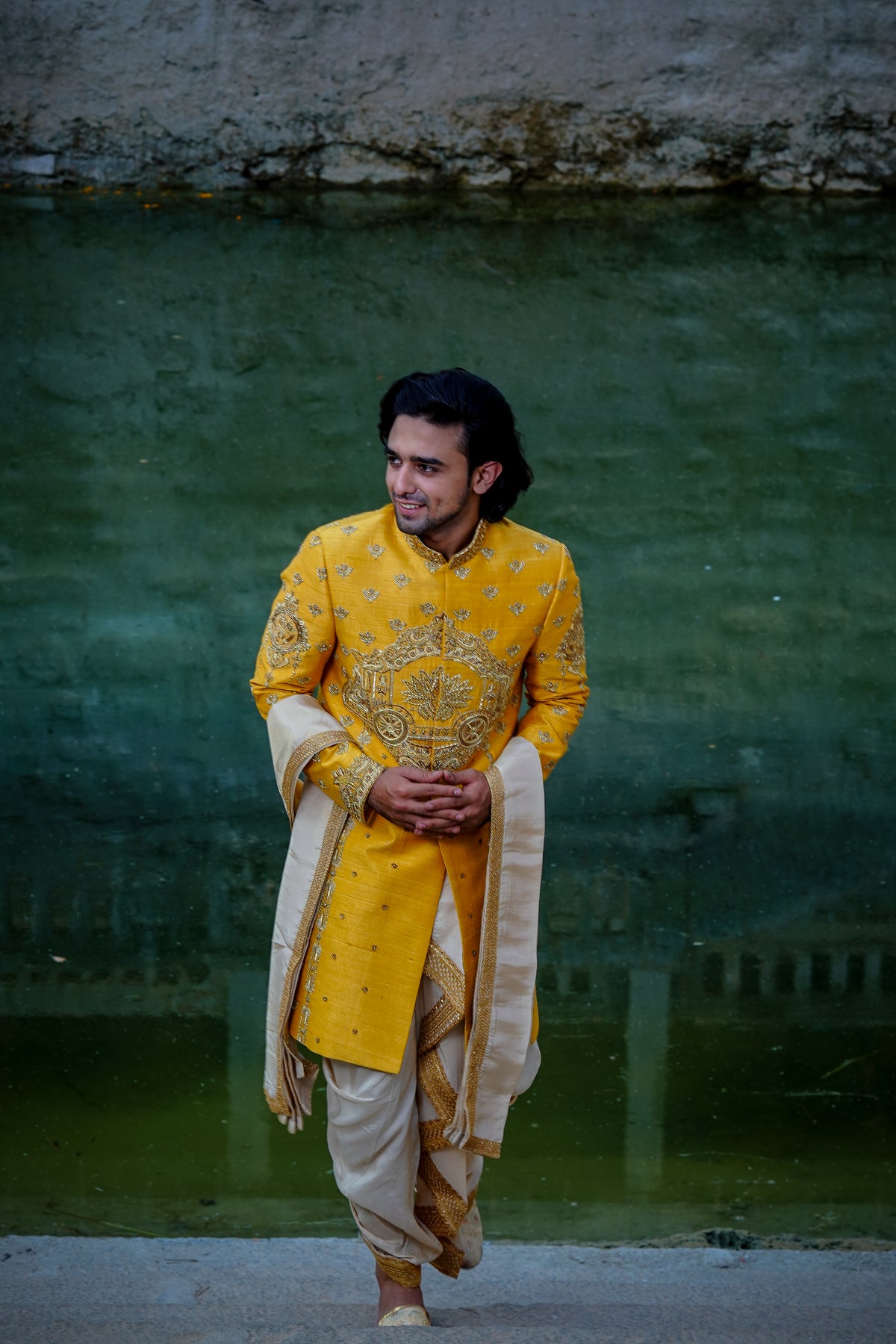 A man wearing a yellow sherwani with gold embroidery and a turban, standing in a traditional setting with a textured background.