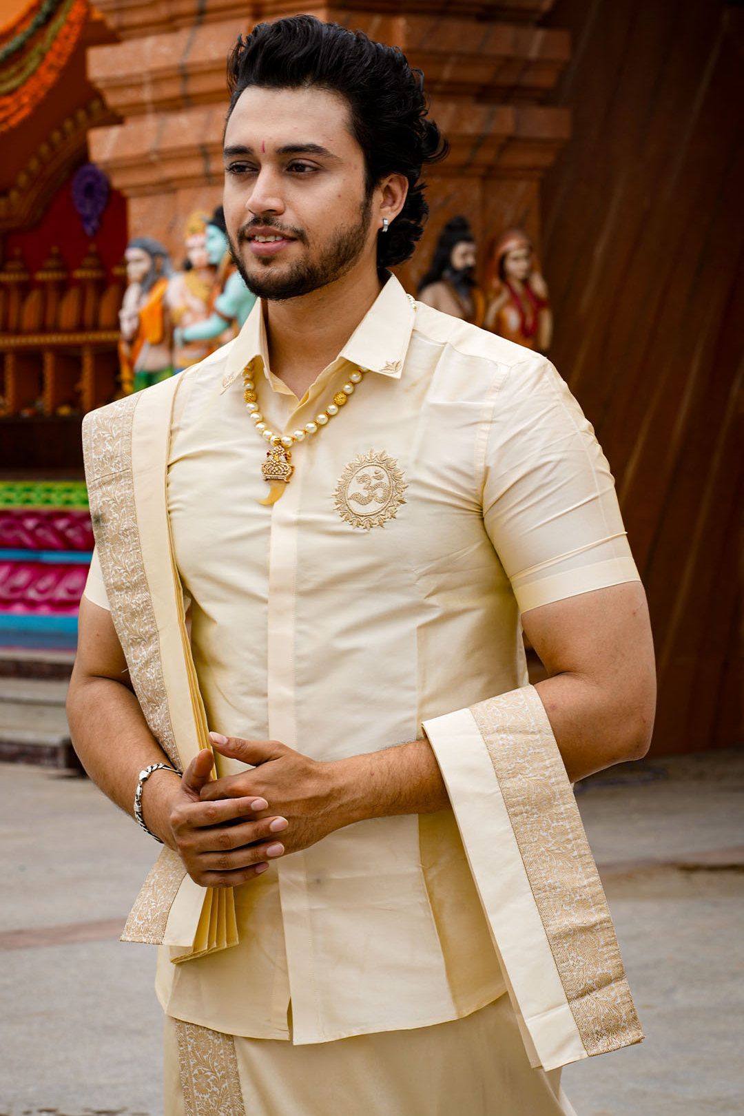 Man wearing a traditional cream-colored outfit with jewelry in an outdoor setting