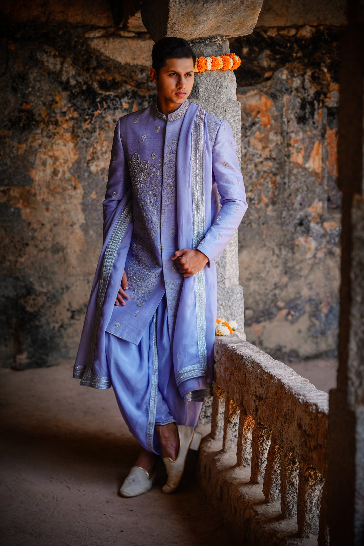 A man wearing a blue Dupam Sherwani with intricate embellishments and opulent embroidery, standing against an outdoor backdrop.