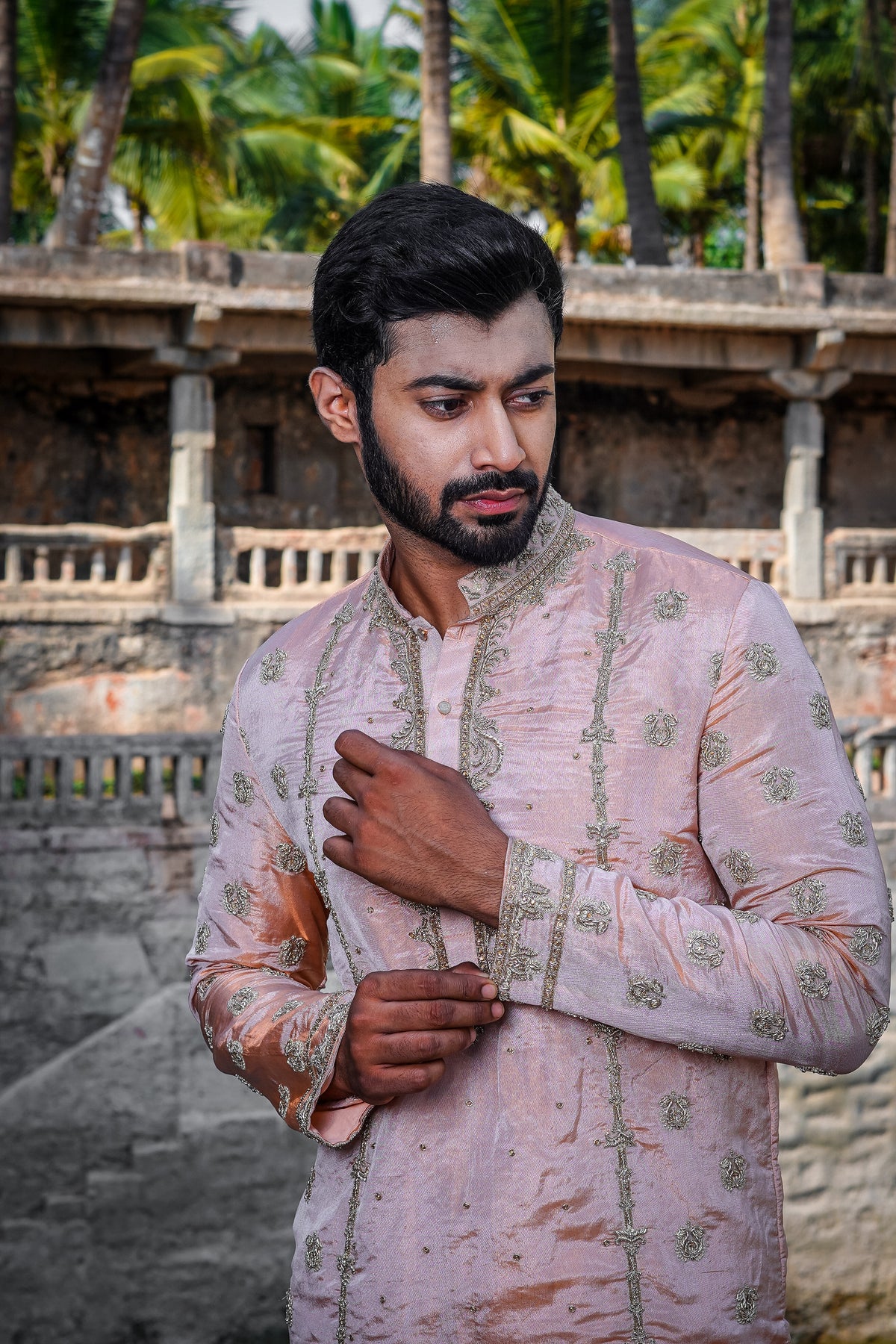 A man standing by a water body, wearing a traditional Sherwani with blue and gold colors, featuring a dupion silk dhoti and khandwa.
