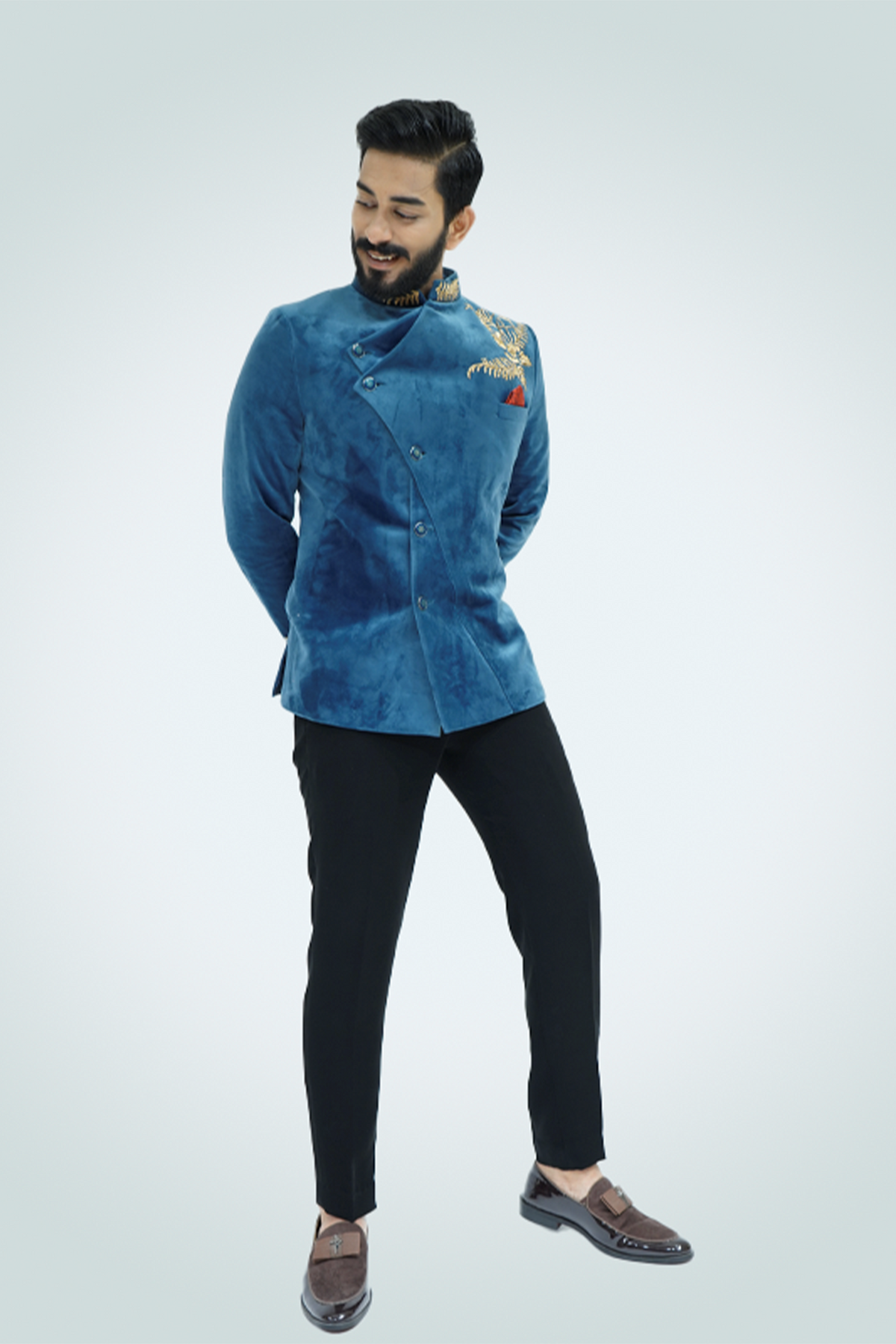 A man wearing a Teal Blue Velvet Jodhpuri with gold embellishments, paired with black trousers and tan shoes.