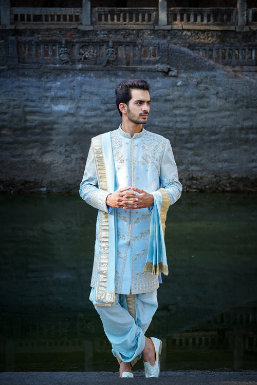 A man standing by a water body, wearing a traditional Sherwani with blue and gold colors, featuring a dupion silk dhoti and khandwa.