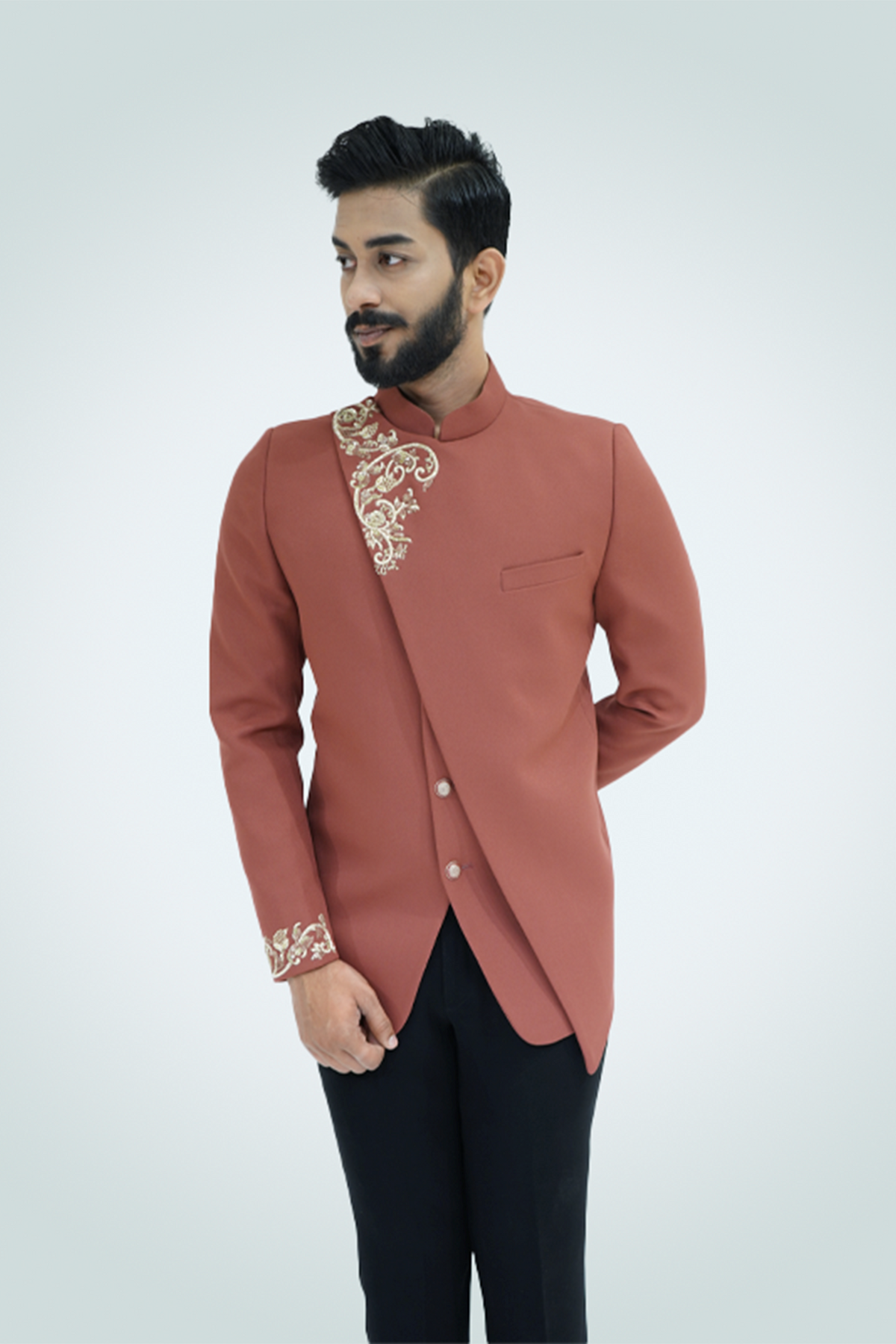 A man wearing a rust-colored Jodhpuri suit with gold embroidery, featuring a mandarin collar and cross cut design.