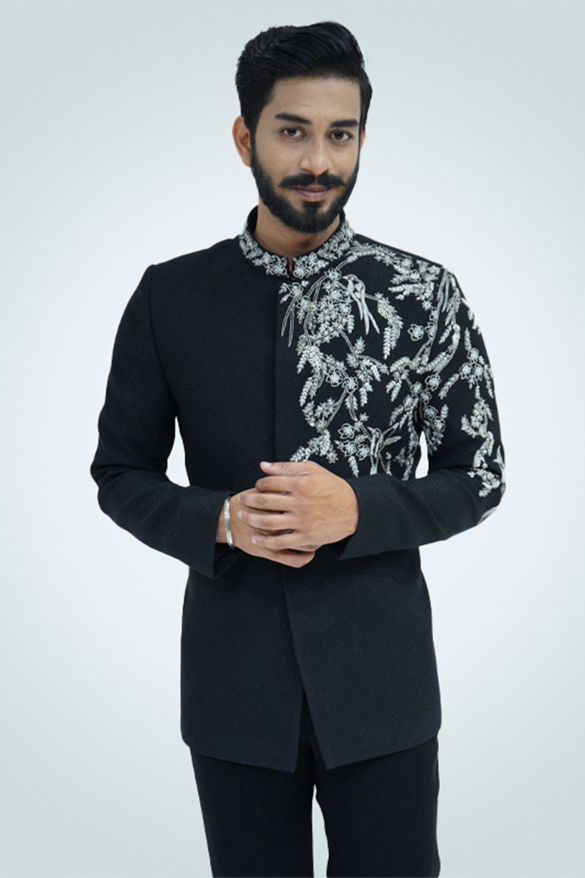 A man wearing a black suit with silver embroidery standing against a light background.