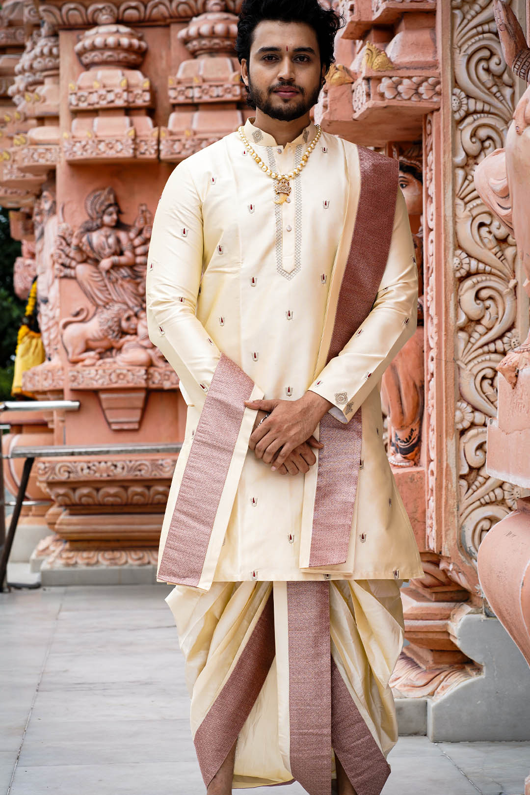 Man in traditional cream and maroon outfit standing in front of ornate pink architectural background