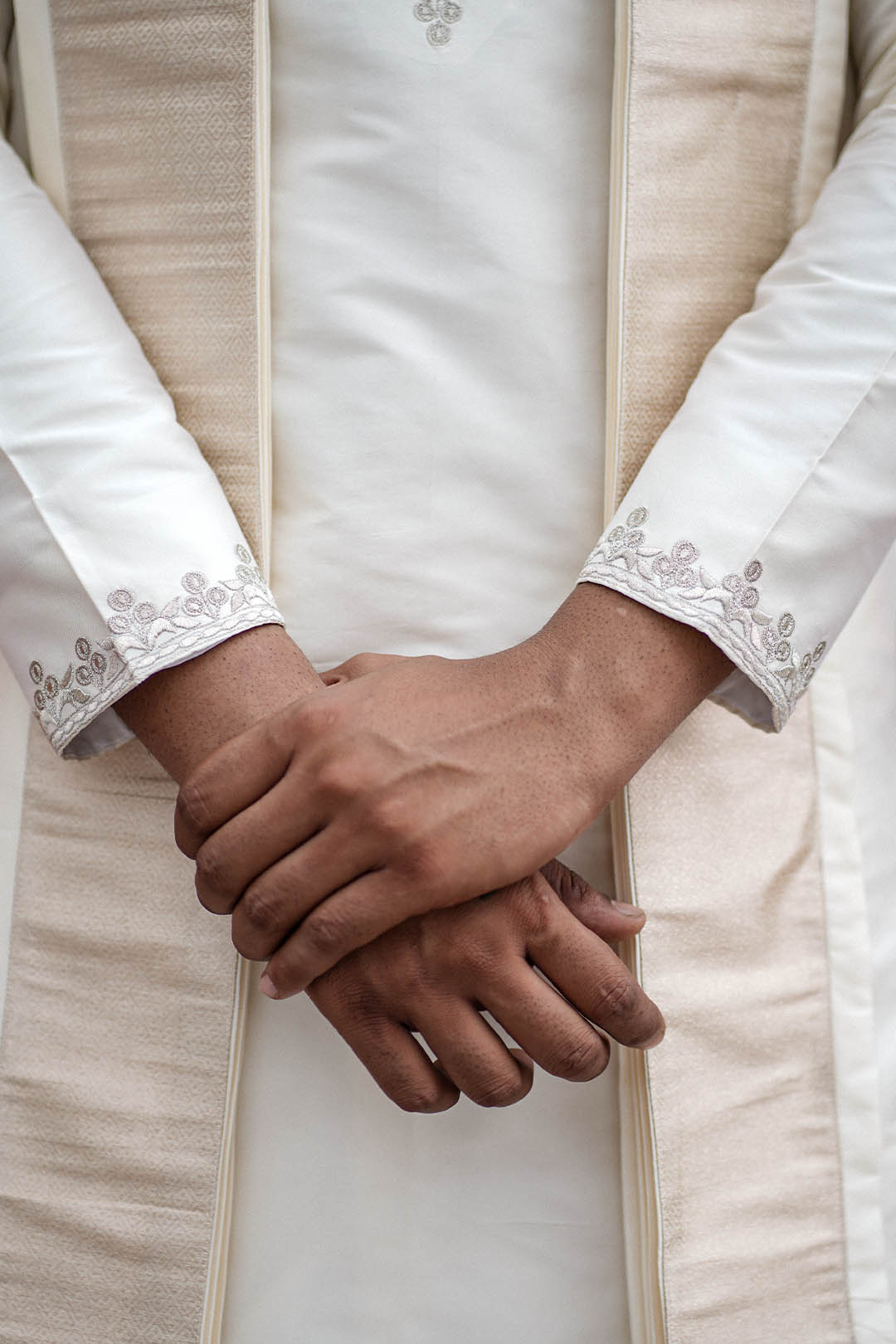 Close-up of hands held together wearing a beige traditional outfit with intricate patterns.