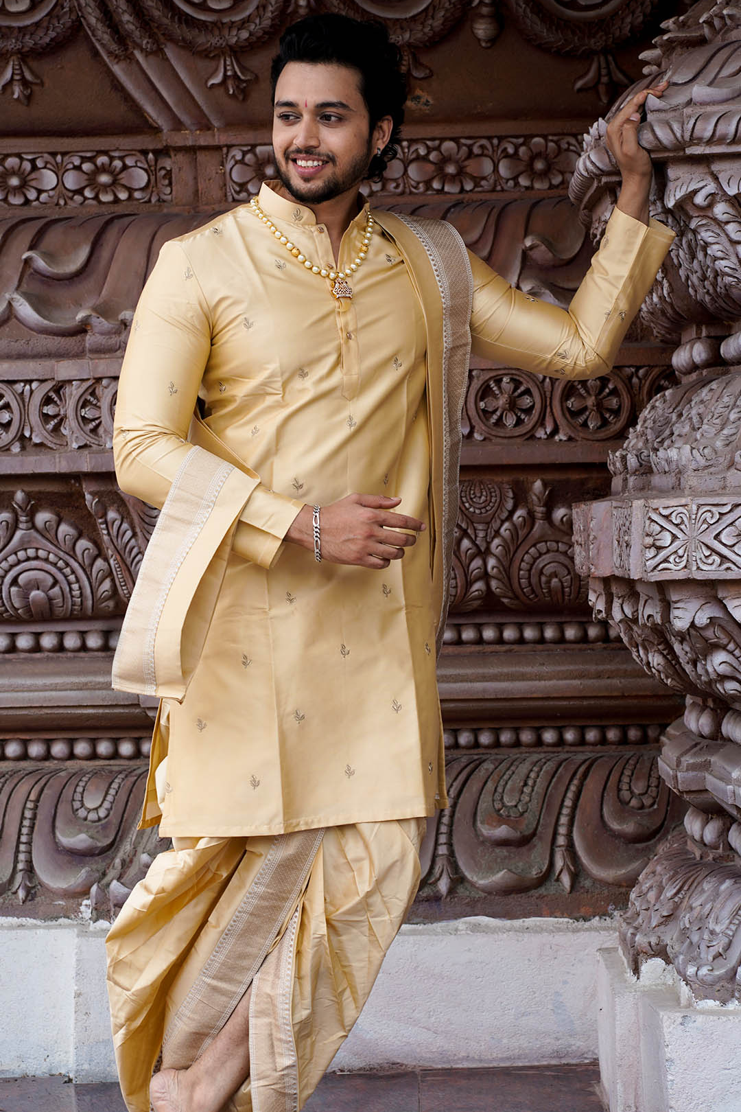 Man in a traditional cream-colored sherwani standing against an ornate architectural background