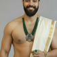 Man wearing a white saree with gold border and green jewelry on a gray background