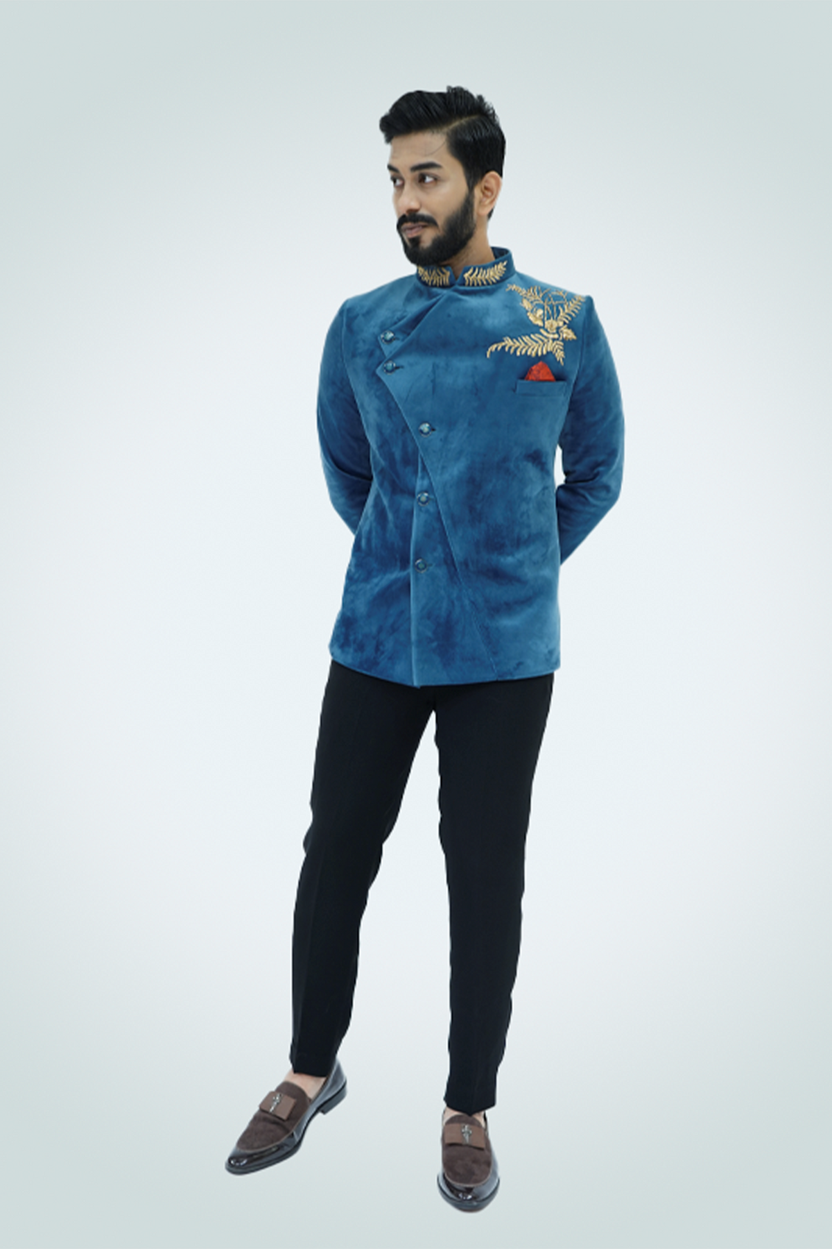 A man wearing a Teal Blue Velvet Jodhpuri with gold embellishments, paired with black trousers and tan shoes.