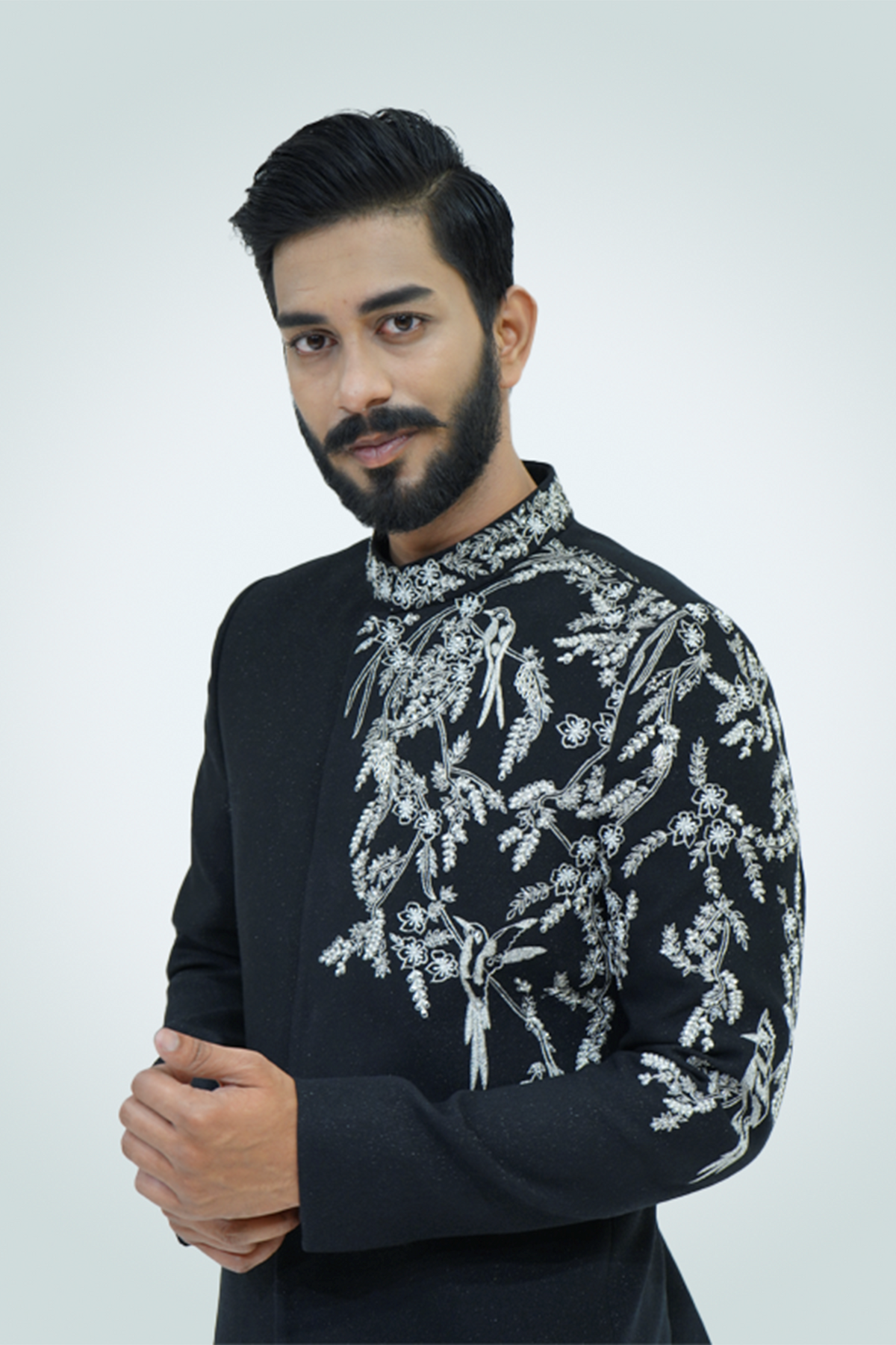 A man wearing a black suit with silver embroidery standing against a light background.