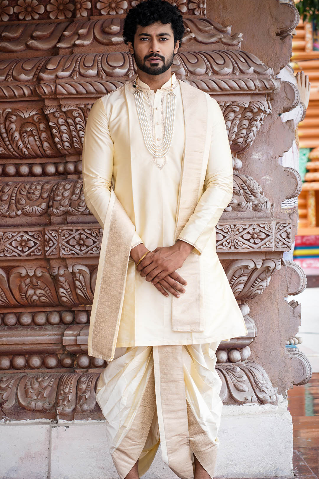 Man wearing a cream traditional outfit standing in front of an ornate stone wall.