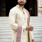Man in traditional cream sherwani with red and gold details standing on steps.