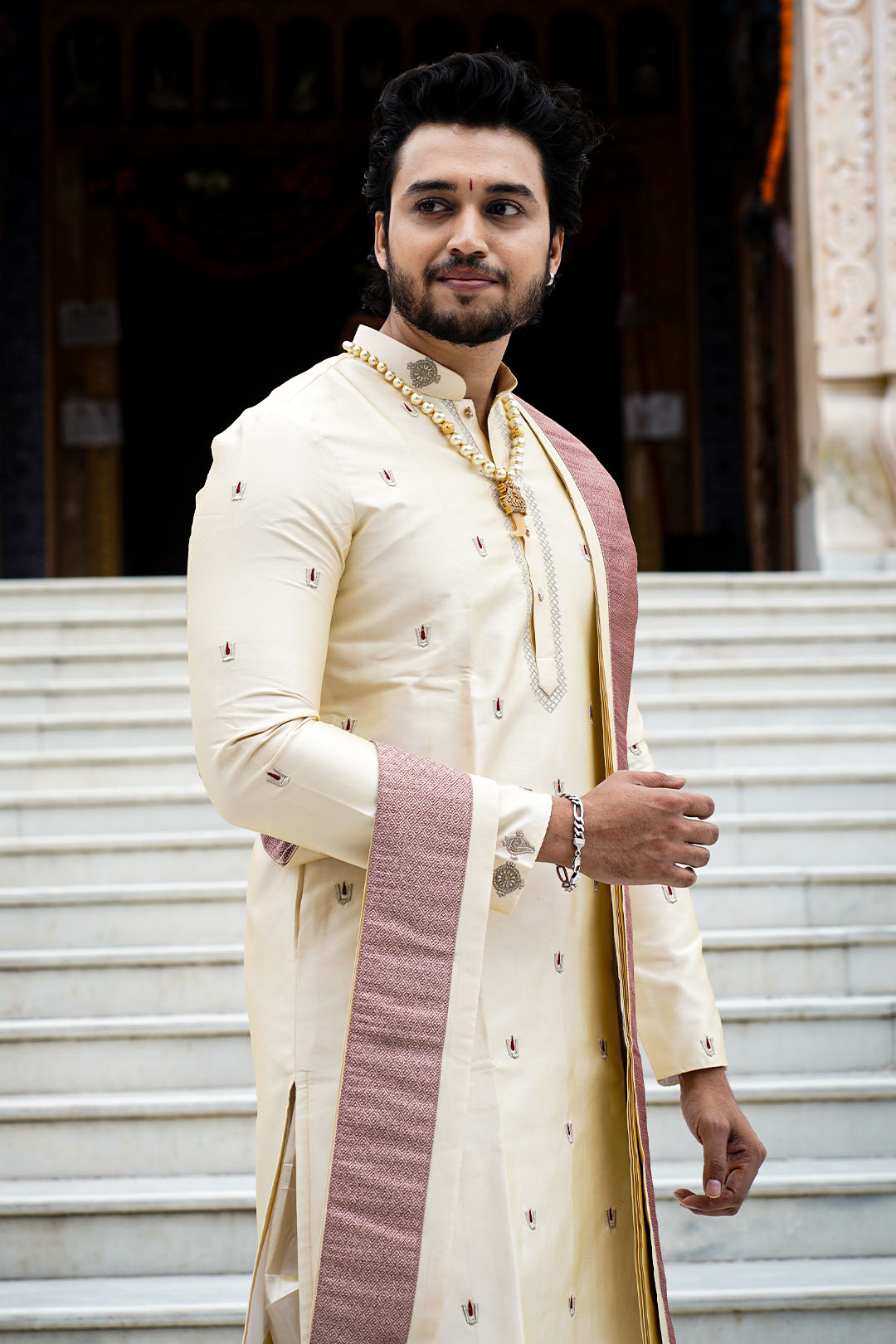 Man in traditional cream sherwani with red and gold details standing on steps.