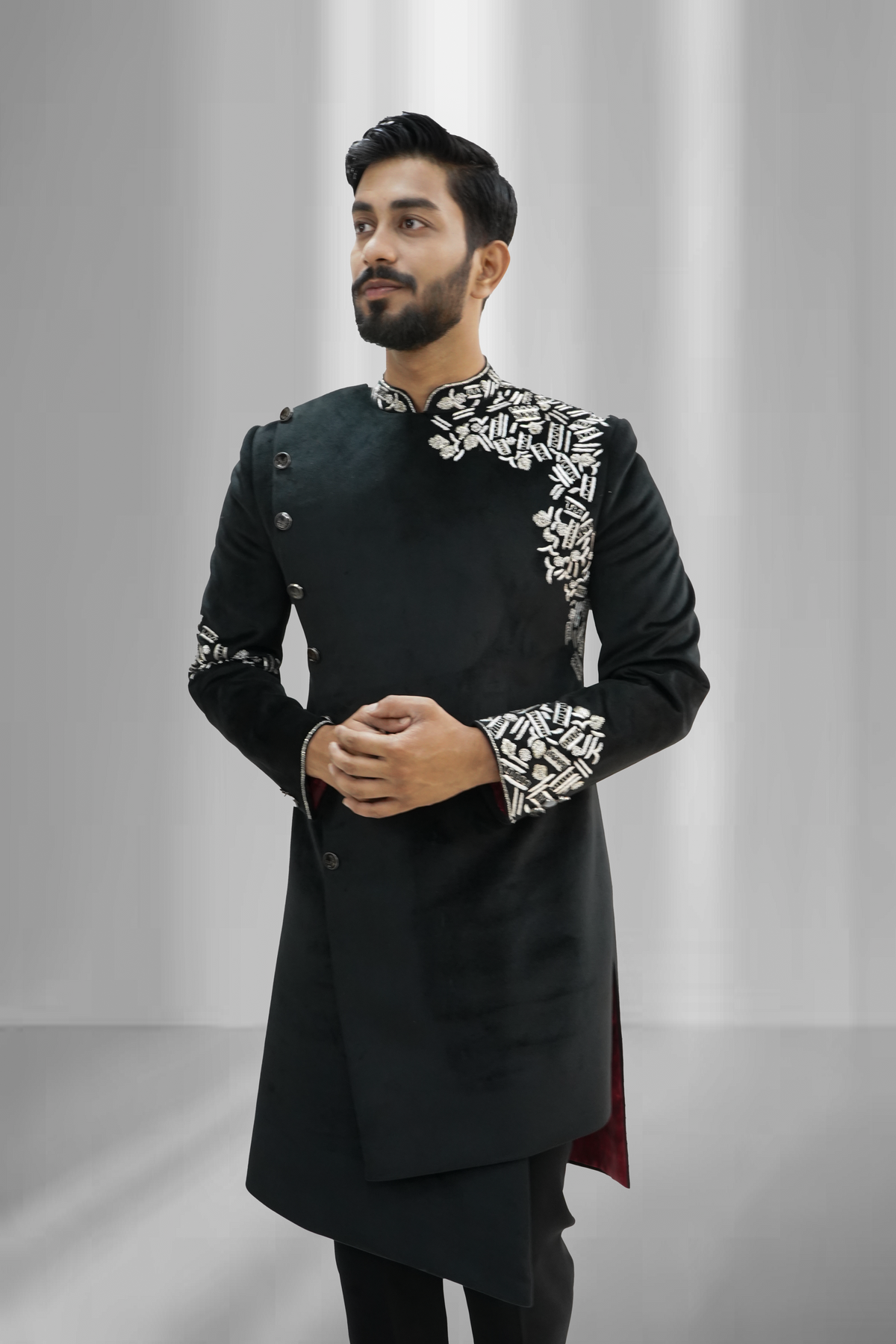 A man wearing a black asymmetrical Indo-Western sherwani with silver zardozi work on the sleeves and chest.