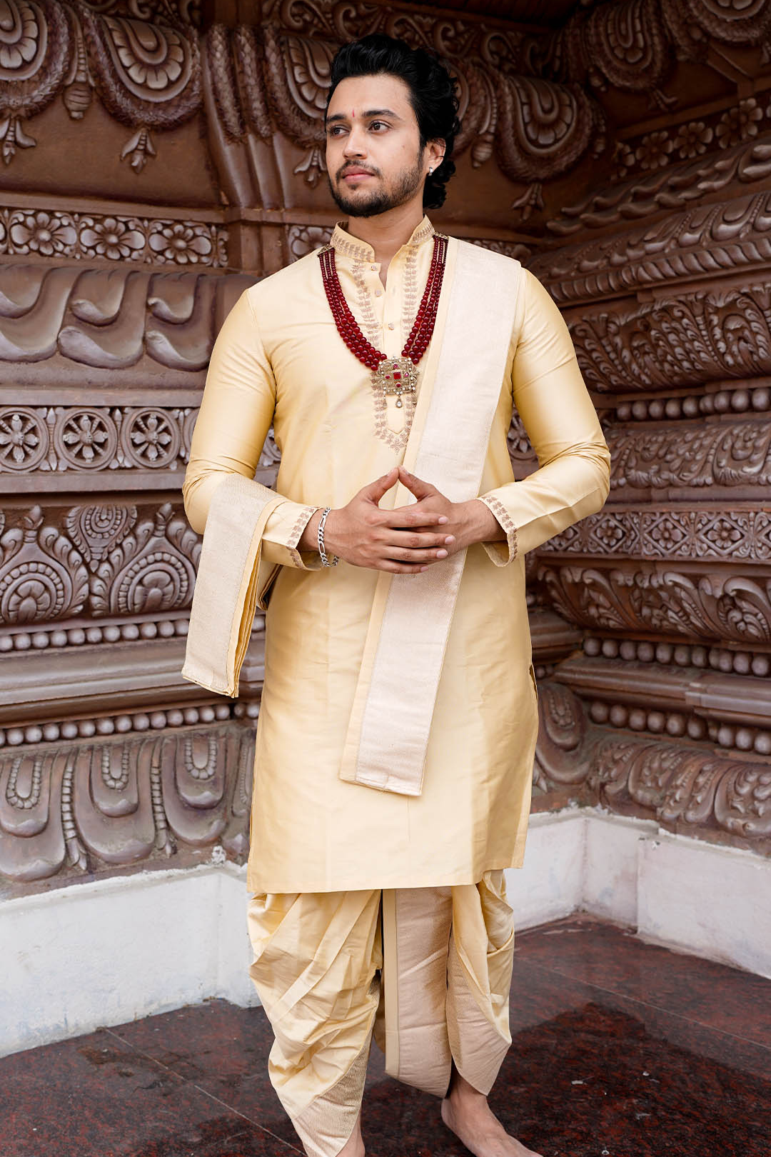 Man in traditional cream kurta with maroon border standing against an ornate brown wall.