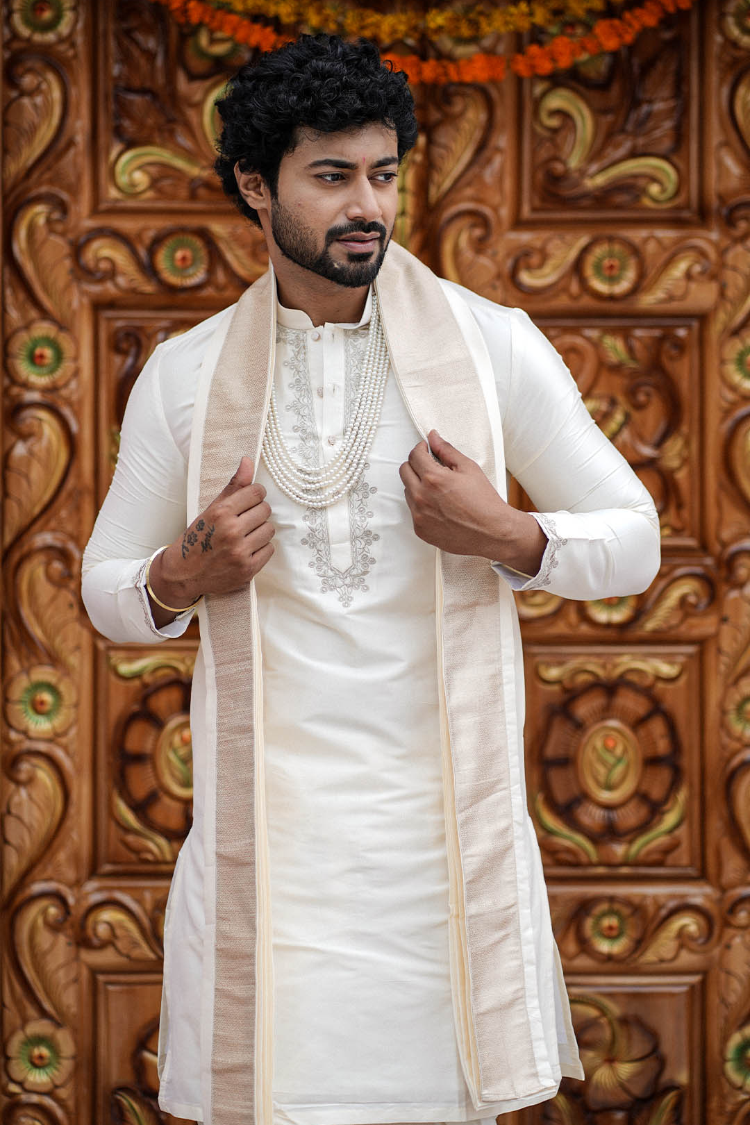 Man wearing a traditional white kurta with a cream shawl against an ornate wooden background