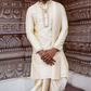 Man wearing a traditional cream-colored sherwani with a gold border against an ornate wall.