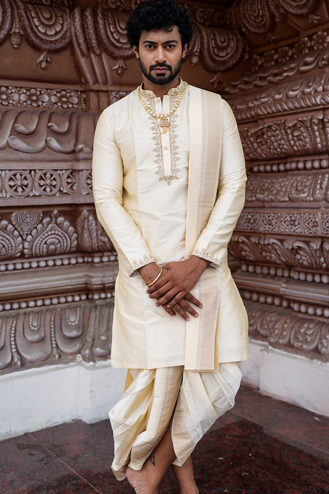 Man wearing a traditional cream-colored sherwani with a gold border against an ornate wall.