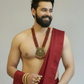 Man wearing a red dhoti with gold jewelry against a plain background