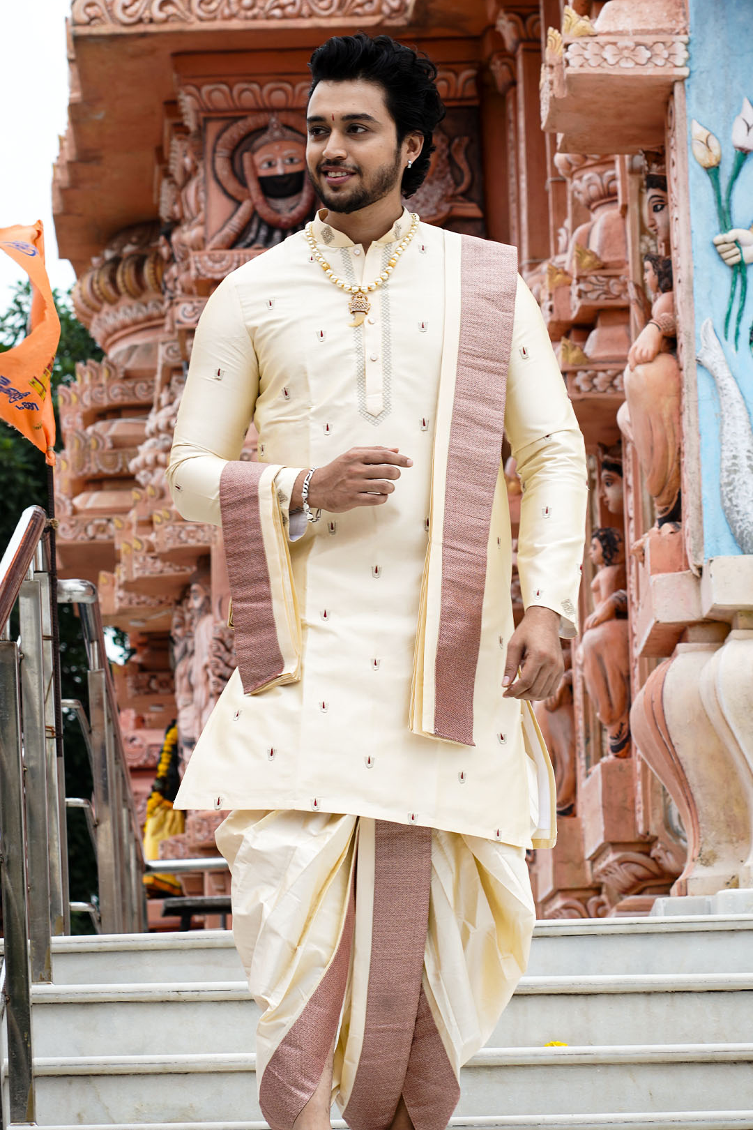 Man in traditional cream and maroon outfit standing in front of a temple.