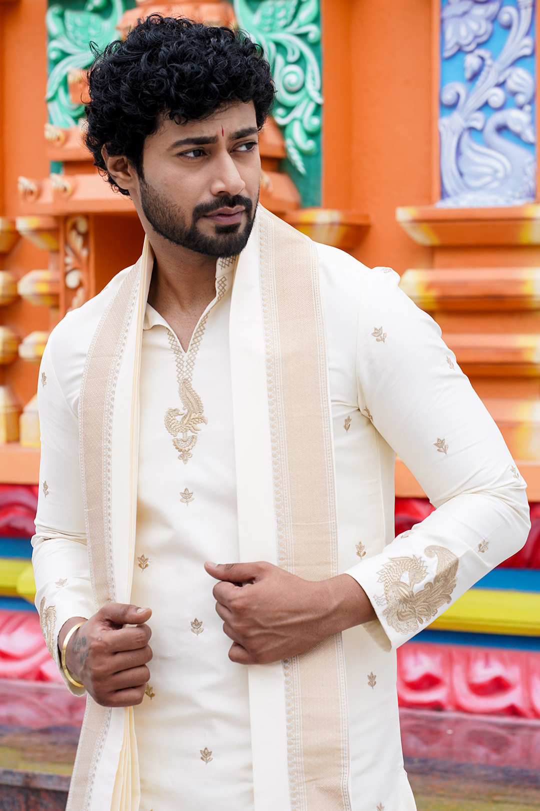 Man wearing a traditional white sherwani with intricate designs against a colorful background